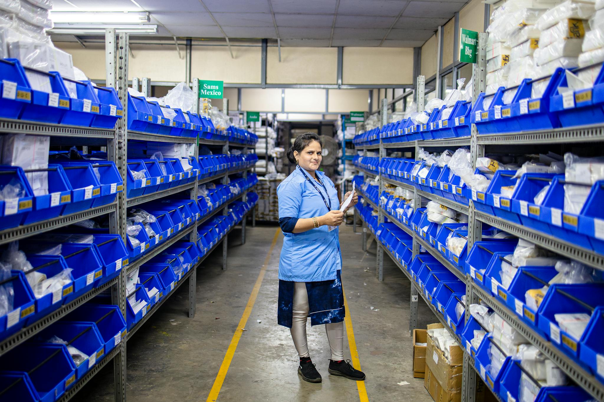 Female worker organizing inventory in a modern warehouse aisle, surrounded by blue storage bins.