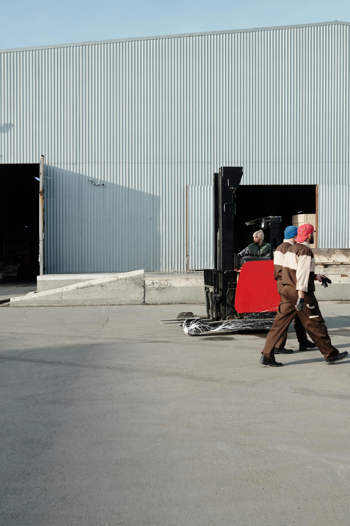 Two workers, one operating a forklift, move goods outside a large warehouse building.